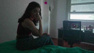 Dalgın genç bir kadın derin düşüncelere dalmış bir ifadeyle yatağın yanında oturuyor. 30 'lu yaşlardaki kadın iç duygularla boğuşuyor.