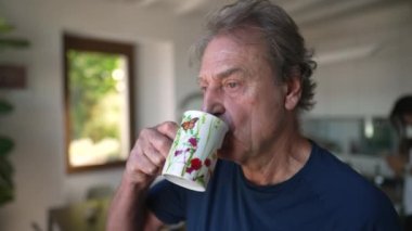 One senior man standing by window drinking tea or coffee in the morning. A pensive older person in 70s looking out in contemplation