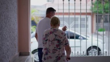 Yetişkin oğlu, yaşlı annesiyle birlikte yürüyüşe çıkmak için evden ayrılıyor, adam kapıyı itiyor ve yaşlı kadının kaldırıma doğru yürümesine yardım ediyor, yaşlı insan hareketliliği.