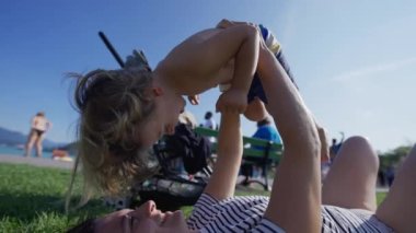 Happy mother bonding with little son laying on grass outside at park. Family lifestyle moment of parent holding child in the air lies outdoors in sunny day