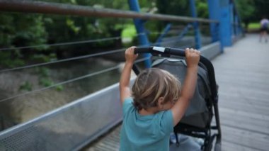 Adorable small boy helping to push stroller outside at park. Family vacations concept. Child pushing baby carriage outdoors crossing bridge