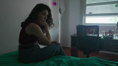 Dalgın genç bir kadın derin düşüncelere dalmış bir ifadeyle yatağın yanında oturuyor. 30 'lu yaşlardaki kadın iç duygularla boğuşuyor.