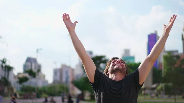 stock image Faithful young man raises arms to sky feeling GRATEFUL standing outdoors in urban city. A male person celebrates LIFE with HOPE