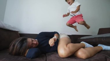 Aktif bir çocuk kanepede annesinin üzerine atlıyor. Mutlu bir çocuk evde eğleniyor.