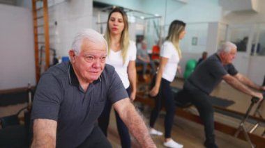Rehberli Egzersiz 'de Kıdemli Adam, Kadın Koçla Pilates Stüdyosu, Esneme ve omurga sağlığıyla ilgilenmek için makineyi kullanan yaşlı kişi