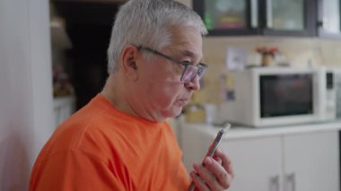 Kıdemli adam telefonda konuşuyor ve sesli mesaj gönderiyor. 70 'li yaşlarda, modern teknolojiyi kullanarak mutfakta yaşlı bir erkek.