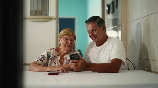 stock image Candid middle-aged son helping elderly mother with smartphone device in home kitchen table. Man assisting old woman with modern technology