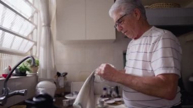 Emekli kıdemli bir adam mutfakta pencerenin önünde dururken çay fincanlarını havluyla kurutuyor. Yaşlı kişi ev yaşamında faaliyet gösteriyor