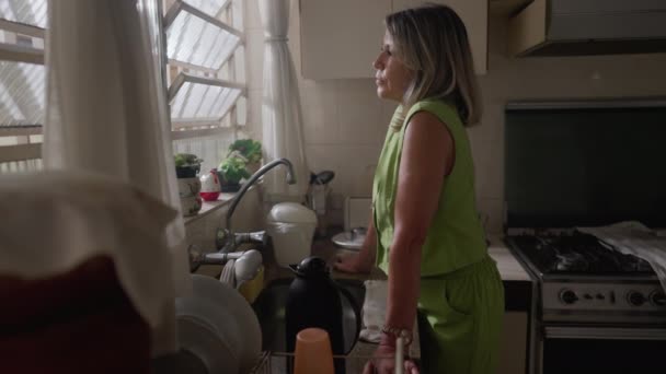 瞑想的な表情でキッチンウィンドウに立っている普及した中年の女性 50代の思慮深い女性は自宅での解決策を熟考 — ストック動画