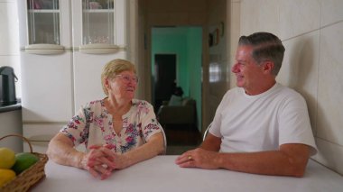 Kıdemli anne, sıradan bir aile hayatı sahnesinde yetişkin oğluyla konuşurken gülümsüyor ve gülüyor. Gerçek hayattaki insanlar ev mutfağında iletişim halindeler.