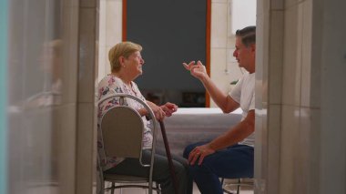 Evde yaşlı bir anneyle konuşan orta yaşlı bir çocuğun gizli aile içi sahnesi. Otantik gerçek hayat, yaşlılık yaşam tarzı, mutfakta oturan yaşlılarla ilgilenen oğul.