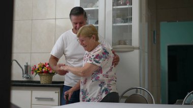 Orta yaşlı bir çocuğun, yaşlı bir anneye sandalyesinden kalkması için yardım ettiği gerçek hayatta yaşlılara yardım eden aile tasviri.