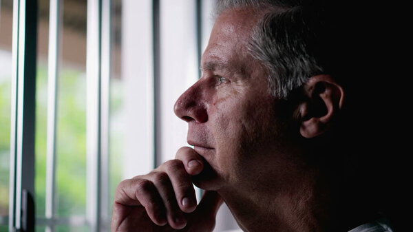 Pensive Middle-Aged Man with Gray Hair Looking Out Window, Contemplative Expression Suggesting Deep Thought about Life, person remembering past memories with hand in chin