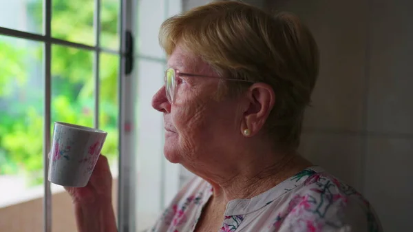 80多岁的老年妇女喜欢喝咖啡或Tea 带着沉思的表情凝视着厨房的窗户 多愁善感的老年家庭生活方式晨礼 — 图库照片