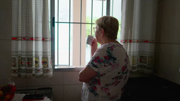 一位80多岁的心烦意乱的老妇人一边喝着咖啡 一边从厨房的窗户向外张望 — 图库照片