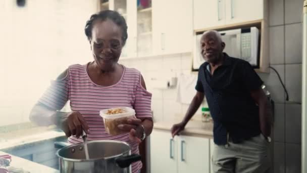 在家里的厨房里 一对快乐的非洲裔美国夫妇 一位老年黑人妻子 与有背景的伴侣站在火炉边 把食物倒入碗里 — 图库视频影像