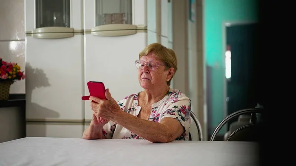 現代技術と関わるおばあちゃん 高齢家庭の台所でスマートフォンデバイスを使用して家庭生活 電話画面で主演 — ストック写真