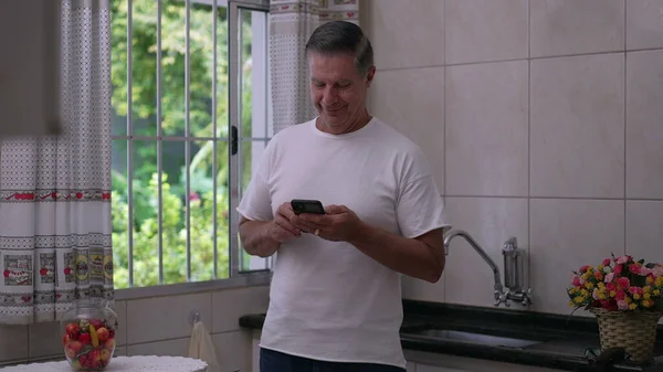 一位中年男子站在自家厨房的窗边 看着智能手机的显示屏 男性50人 在家庭休闲生活中使用现代技术 — 图库照片