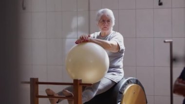Pilates sınıfındaki düşünceli son sınıf öğrencisi balo düzenliyor, yaşlı bayan antrenman sırasında dinleniyor.