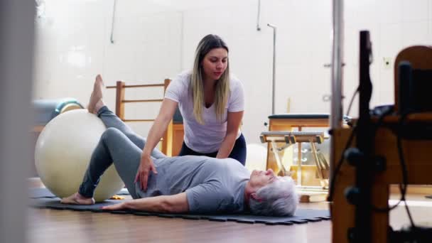 理疗师指导老年妇女用普拉提球进行运动 一位老妇人在一位体育教练的帮助下 在地板上进行日常锻炼 — 图库视频影像