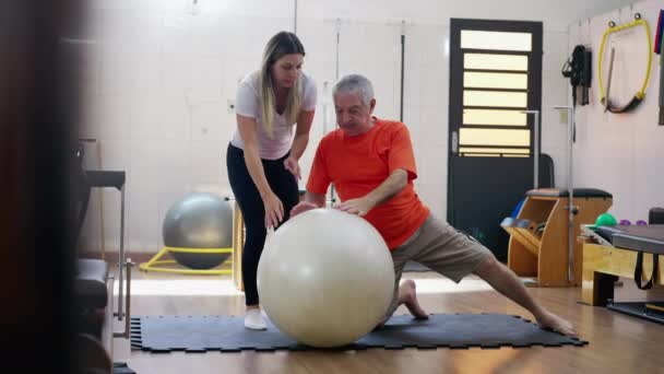 ピラティスインストラクタージムボールで強化と柔軟性の行使で高齢者を支援 — ストック動画