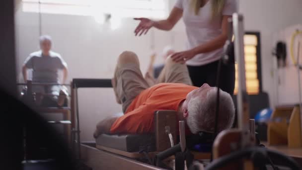 Starszy Mężczyzna Leżał Maszynie Pilates Wzmacniającej Ciało Pomocą Trenerki Pilates — Wideo stockowe