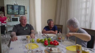 Bir grup yaşlı arkadaş samimi, sıradan bir ev ortamında öğle yemeği için toplandı. Oturma odasında çeşitli yaşlılar konuşuyor.