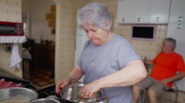 Son sınıf öğrencisi kadın ocağın yanında yemek pişiriyor. Beyaz saçlı, yaşlı, beyaz bir kadın. Karışık yemek.