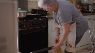 Kıdemli kadın fırını açıyor, yemek hazırlıyor. Önlük giymiş samimi, otantik yaşlı bir büyükanne.