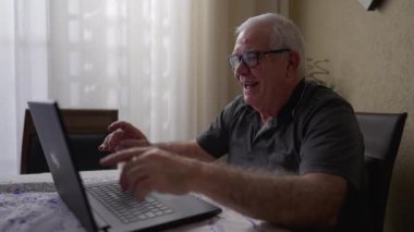 Evde bilgisayarını kullanan mutlu teknoloji meraklısı kıdemli bir adam komik online içeriğe tepki veriyor. Modern teknolojiyle uğraşan yaşlı bir beyaz erkek.