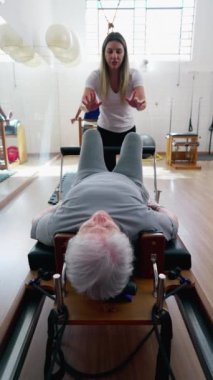 Kadın pilates eğitmeni, yaşlı bir kadına güç makinesiyle egzersiz yaptırıyor. Fizyoterapist stüdyosunda yaşlılık egzersizleri.