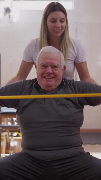 老年男子在一名普拉提女教练的帮助下弯曲橡皮筋进行锻炼 在垂直录像中指导老年男性高加索人锻炼的妇女 — 图库视频影像
