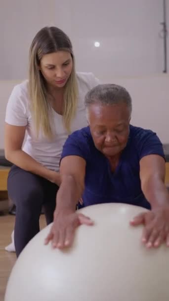 普拉提教师帮助老年黑人妇女进行球类运动 老年人个人锻炼的灵活性和日常锻炼的强度 垂直录像 — 图库视频影像