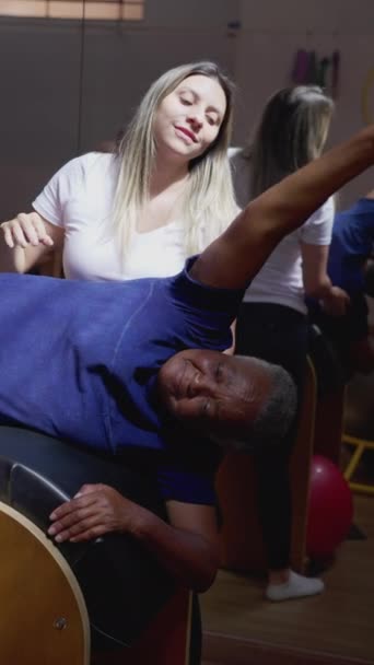 ピラティスのコーチは 理学療法士のセッション中に行使するためにシニア黒人女性を助ける 地元のサービススポーツ事業の女性の所有者は トレーニングに人を支援 — ストック動画