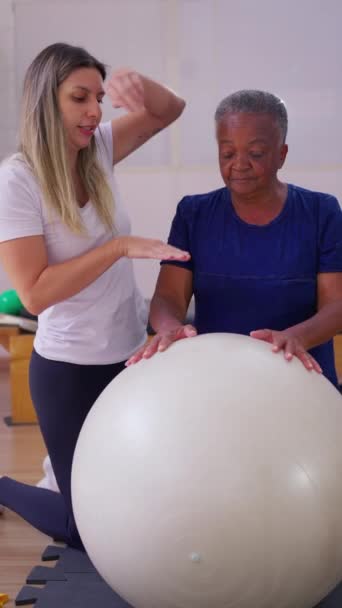 帮助一名巴西黑人老年妇女参加普拉提球运动的女理疗师 垂直录像 — 图库视频影像