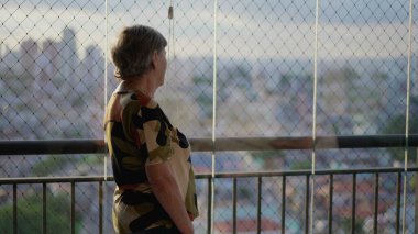 Yaşlı, emekli bir kadın apartmanın balkonunda durmuş şehir manzarasına bakıyor. Şehir manzarasına bakan 70 'li yaşlarda yaşlı bir kadın.