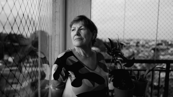 忧心忡忡的老妇人孤身一人站在窗边 凝视着城市的景色 陷入了深深的沉思 穿着单色黑白相间的老年退休妇女 — 图库照片