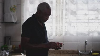 Mutfak penceresinin yanında telefonu kullanan siyahi bir ihtiyar. Bir Afrikalı Amerikalı yaşlının evdeki akıllı telefonuyla online içeriği kontrol ettiği otantik ev sahnesi
