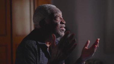 Kıdemli Afro-Amerikan Adam Meditasyonda diz çöküp evde dua ediyor, ellerini kaldırıp yardım istiyor, Tanrı 'ya tapıyor.