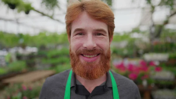 Portrait of a happy redhead male worker wearing Green apron standing inside flower shop. Local business concept of man employee