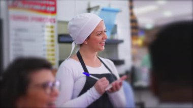Lokantada çalışan mutlu bir garson, müşterilerle konuşurken yemek siparişi alıyor. Yerel işletmelerde geleneksel kadın sunucu mesleği önlük takmak