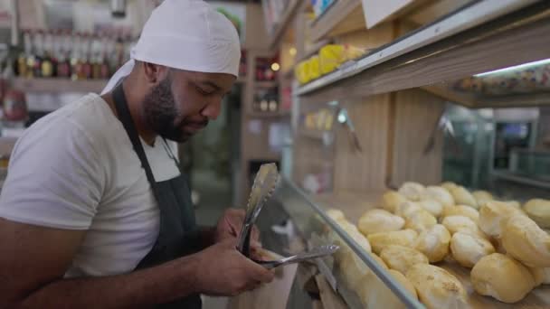 デリカフェテリアで働いている黒人従業員がカウンターの後ろの顧客を支援しています 地元の食料品店でパンを注文する女性 — ストック動画