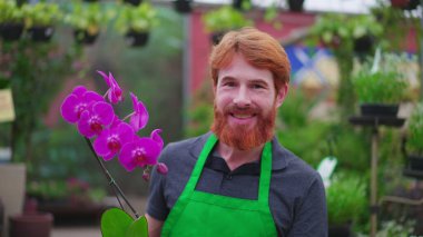Bahçecilik dükkanının neşeli erkek çalışanı elinde çiçeklerle yerel bir dükkanda duruyor. Yeşil önlük giyen kızıl saçlı genç adamın portresi.