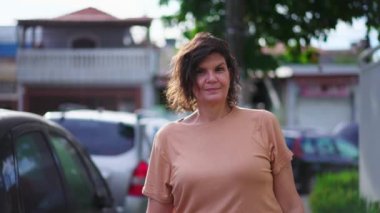 Orta yaşlı bir kadın kaldırımda dikilip kameraya tarafsız bir ifadeyle bakıyor.
