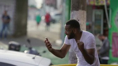 Şaşırmış siyah Brezilyalı bir adam telefondaki uyarıya tepki veriyor. İnançsızlık ve şaşkınlıkla ekranı işaret ediyor. Şehir ortamında sokakta duruyor.