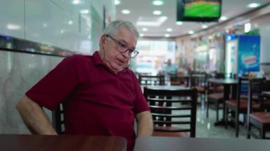Son sınıf öğrencisi okuma gözlüğü takıyor ve cep telefonundan Brezilya kafeteryasının içinde oturuyor. Restoranın içinde modern teknoloji akıllı telefon kullanan yaşlı kişi.