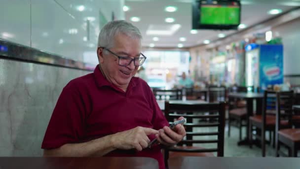 Idoso Feliz Usando Dispositivo Celular Sentado Dentro Cafetaria Brasileira Rindo — Vídeo de Stock