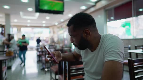 一位巴西黑人男子坐在传统自助餐厅里 用智能手机上网 被在线媒体吸引的人拿着电话盯着屏幕 — 图库视频影像