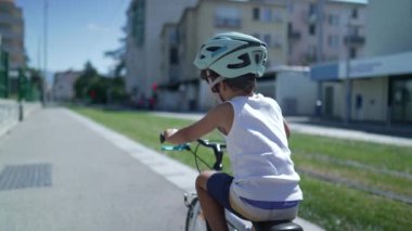 Şehir caddesinde bisiklete binen bir çocuğun arkasında. Kentsel çevre kaldırımında kask takan bisikletli küçük bir çocuk.