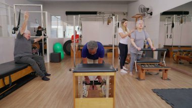 Bir grup yaşlı insan, bayan antrenör eşliğinde pilates seanslarına katılıyor. Yaşlı erkekler ve kadınlar vücut ve egzersiz için makineler kullanıyor, yaşlılık egzersizleri.